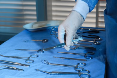 Photo of Doctor taking scalpel from table with different surgical instruments indoors, closeup