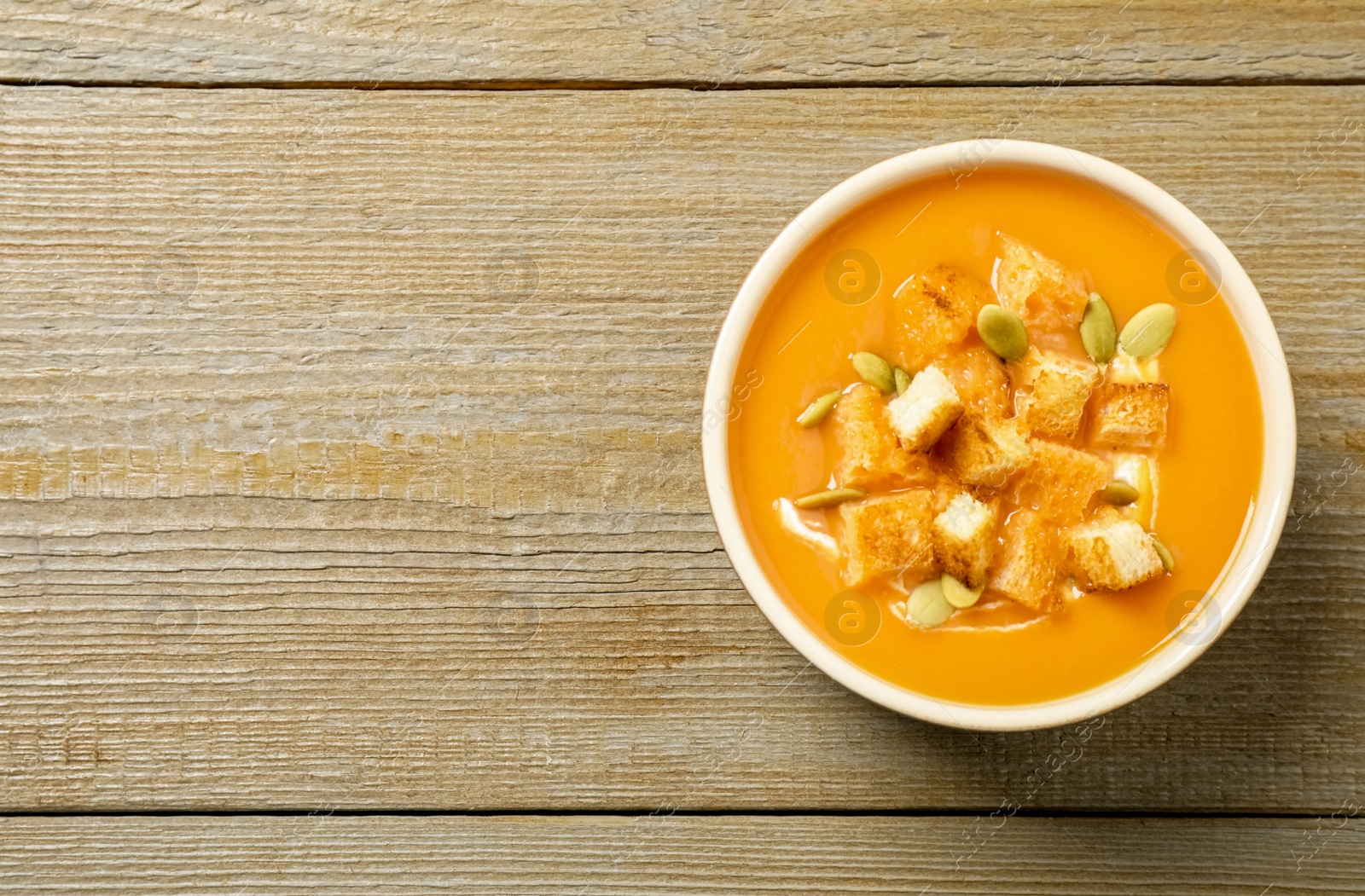 Photo of Tasty creamy pumpkin soup with croutons and seeds in bowl on wooden table, top view. Space for text