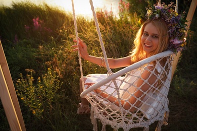 Photo of Young woman wearing wreath made of beautiful flowers on swing chair outdoors at sunset