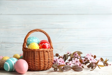 Photo of Painted Easter eggs in wicker basket and blossoming branches on table. Space for text