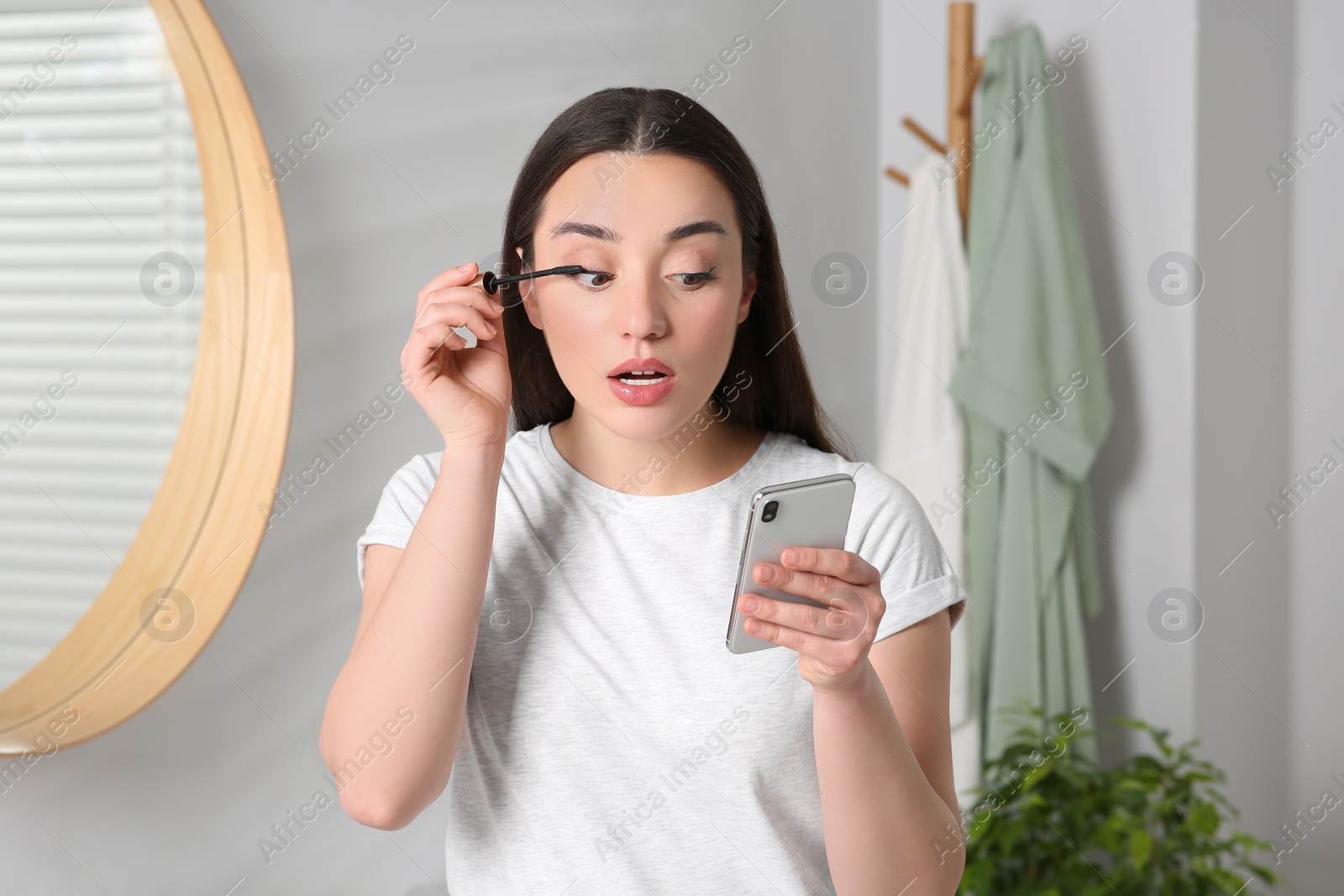 Photo of Beautiful young woman using smartphone while applying mascara in bathroom. Internet addiction