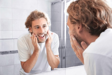 Photo of Young man washing face with soap near mirror in bathroom