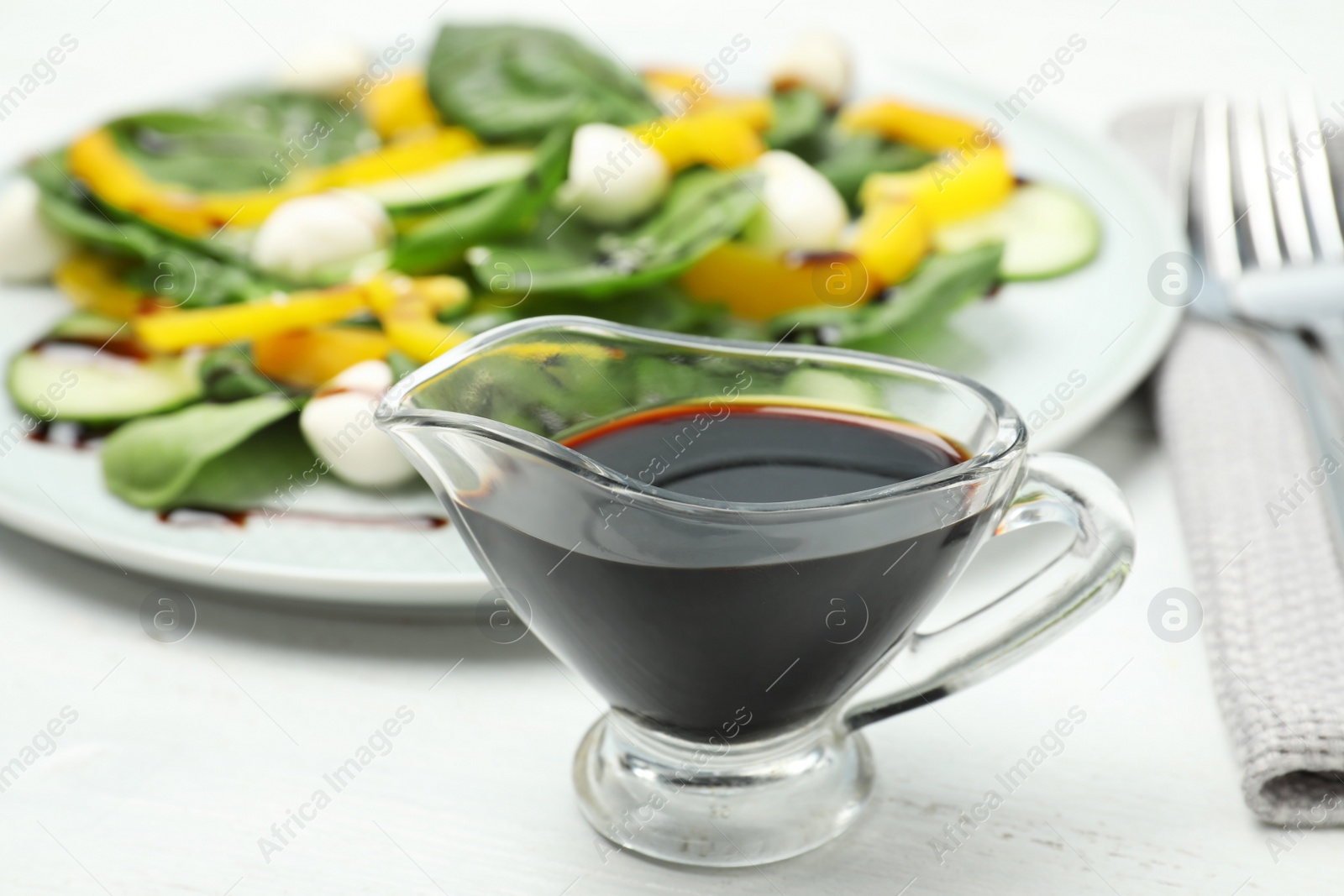 Photo of Balsamic vinegar in glass gravy boat near plate with vegetable salad on table, closeup