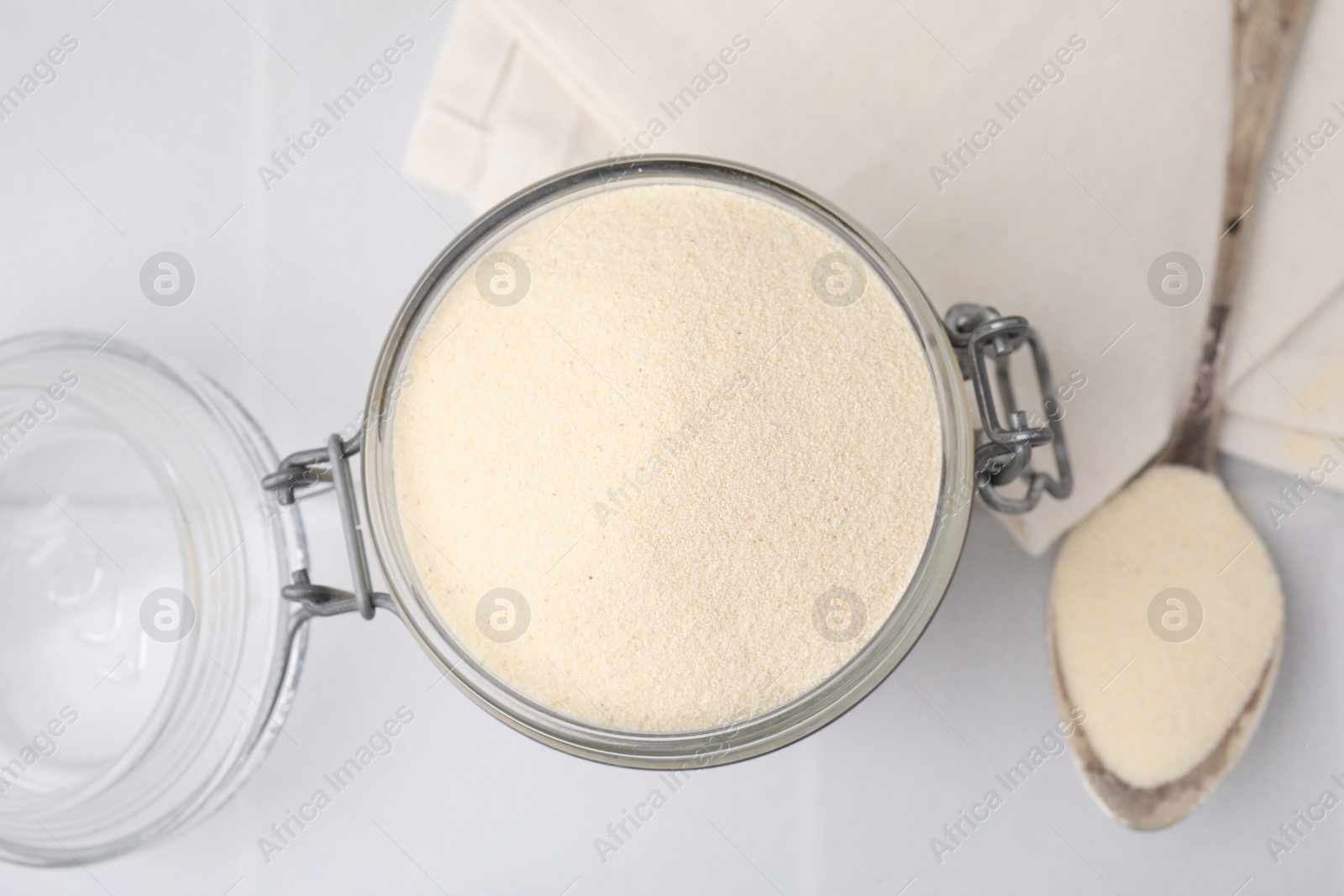 Photo of Uncooked organic semolina in jar and spoon on white tiled table, top view