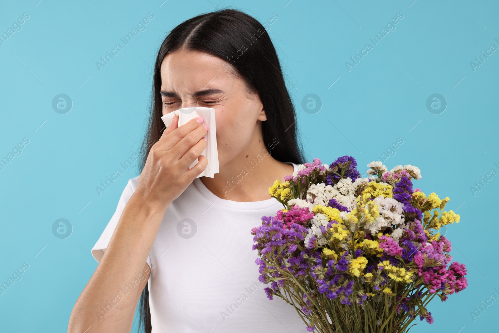 Photo of Suffering from allergy. Young woman with bouquet of flowers blowing her nose in tissue on light blue background