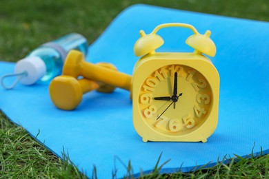 Photo of Alarm clock, dumbbells, bottle of water and fitness mat on green grass outdoors, closeup. Morning exercise