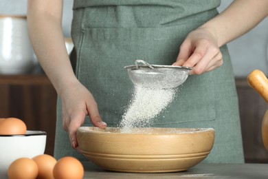 Photo of Woman sieving flour into bowl at table in kitchen, closeup