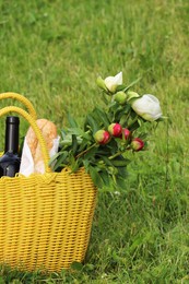 Yellow wicker bag with wine, bread and flowers on green grass outdoors. Picnic season