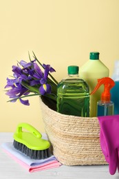 Photo of Spring cleaning. Wicker basket with detergents, flowers and gloves near tools on white wooden table