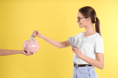 Photo of Young woman putting money into piggy bank on yellow background