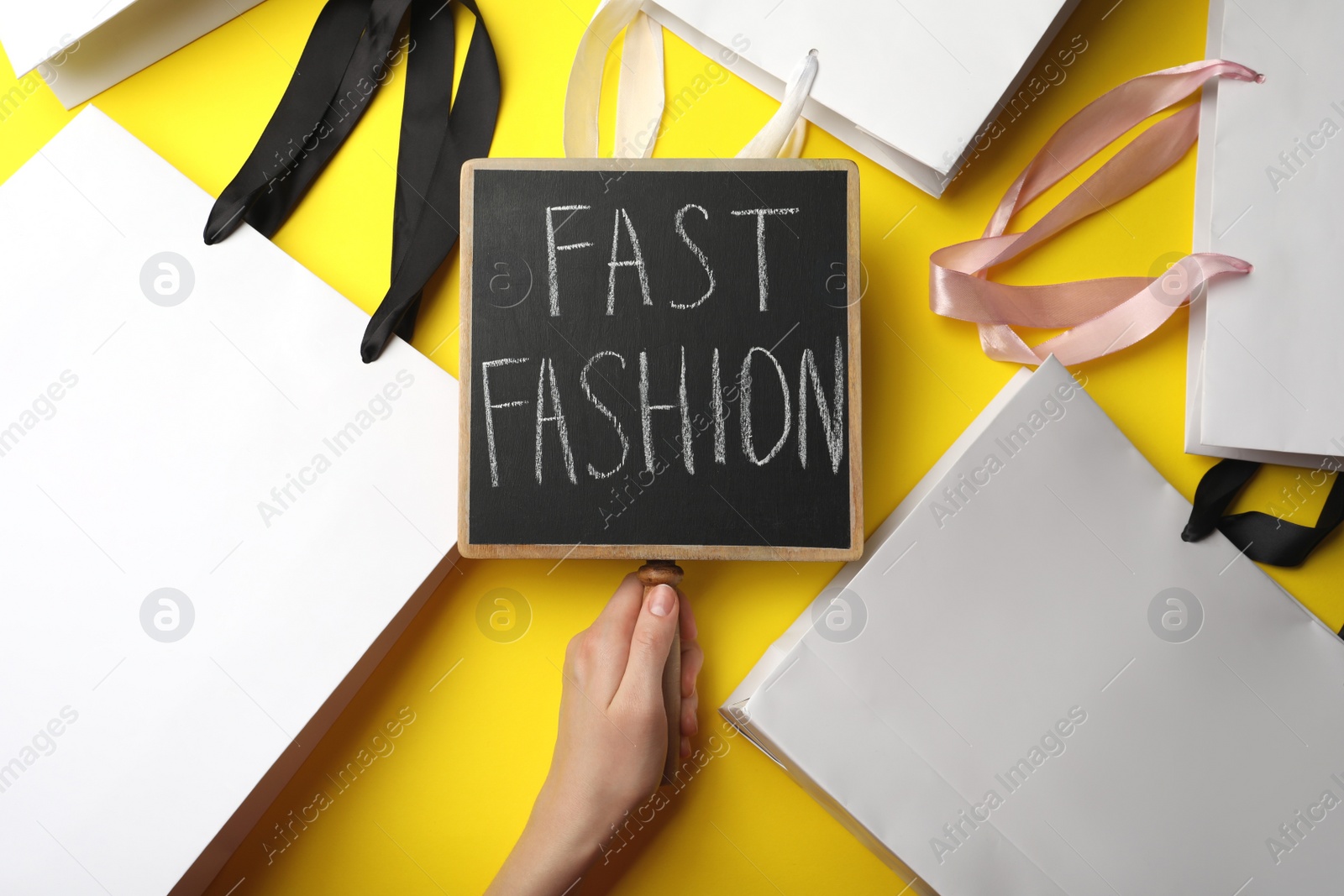 Photo of Woman holding small chalkboard with phrase Fast Fashion and white shopping bags on yellow background, top view