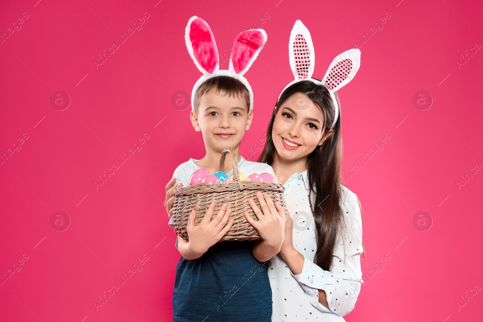 Photo of Mother and son in bunny ears headbands with basket of Easter eggs on color background