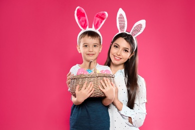 Photo of Mother and son in bunny ears headbands with basket of Easter eggs on color background