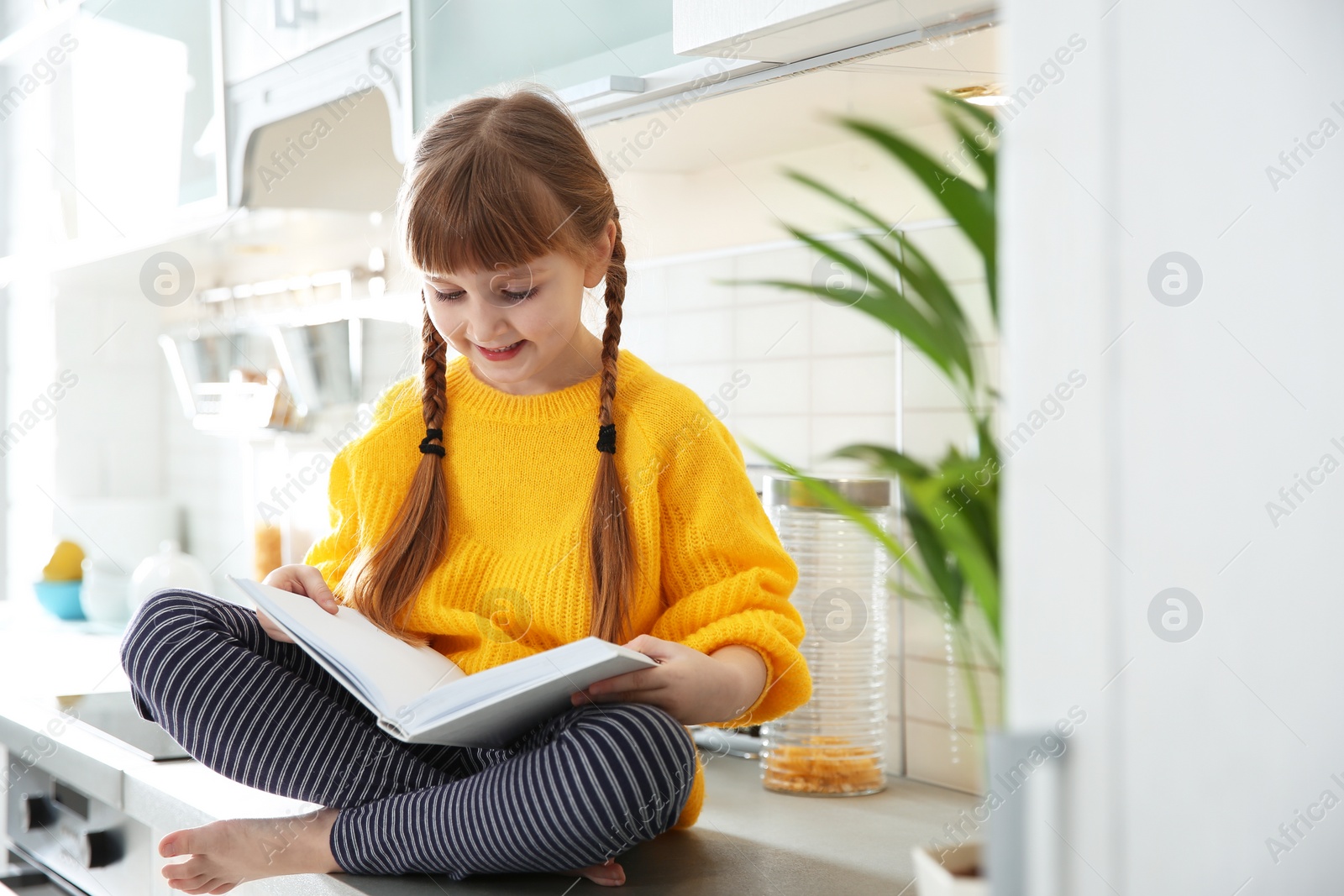 Photo of Cute little girl reading book in kitchen at home