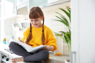 Cute little girl reading book in kitchen at home