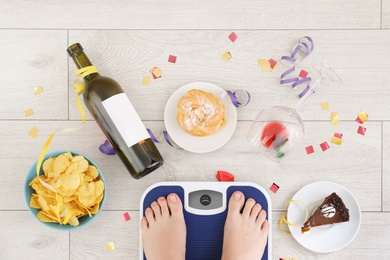 Photo of Woman standing on scales surrounded by different food and alcohol after party indoors, top view