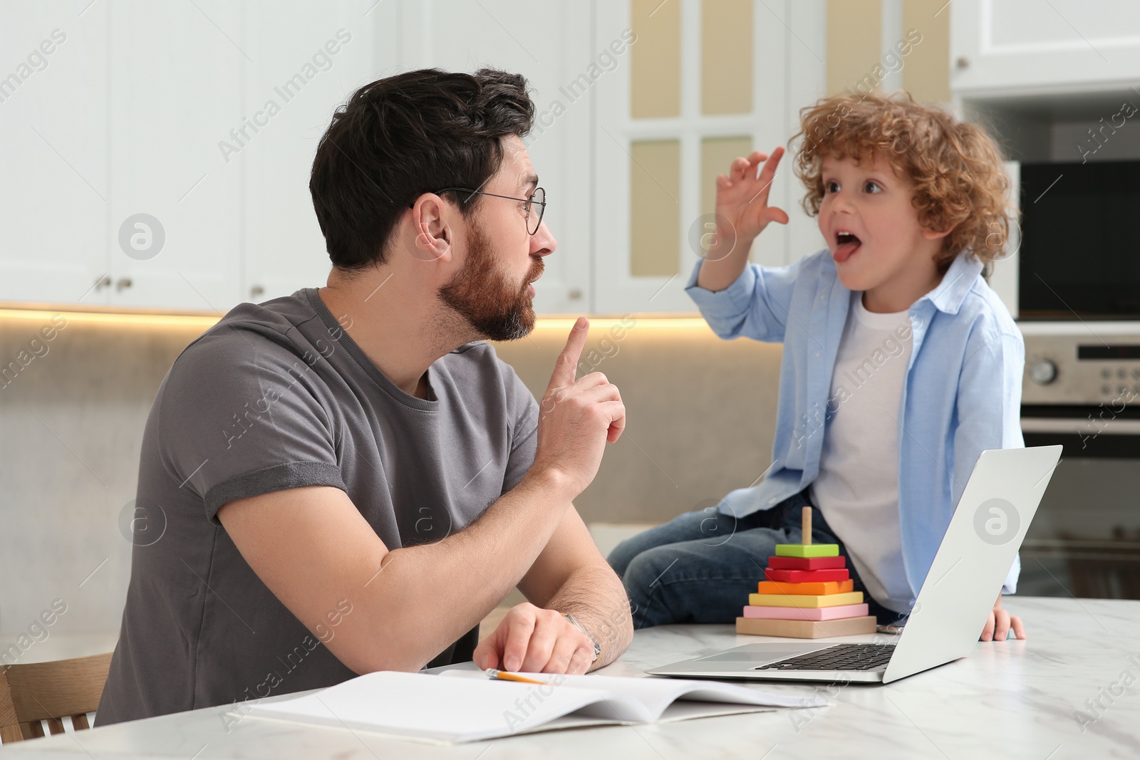 Photo of Little boy bothering father at home. Child playing with toys on desk while his father working remotely