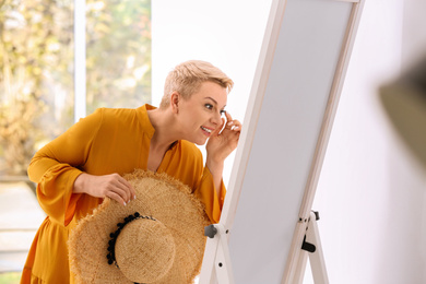 Photo of Beautiful mature woman with hat looking at herself in large mirror indoors