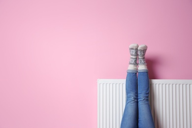 Photo of Woman warming legs on heating radiator near color wall. Space for text