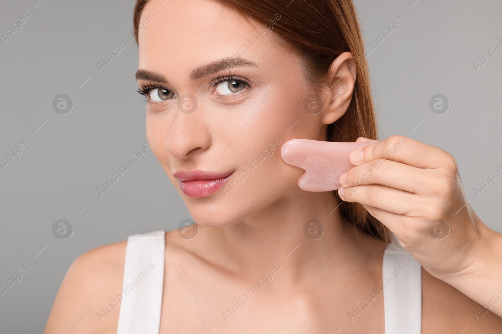Photo of Young woman massaging her face with rose quartz gua sha tool on grey background, closeup