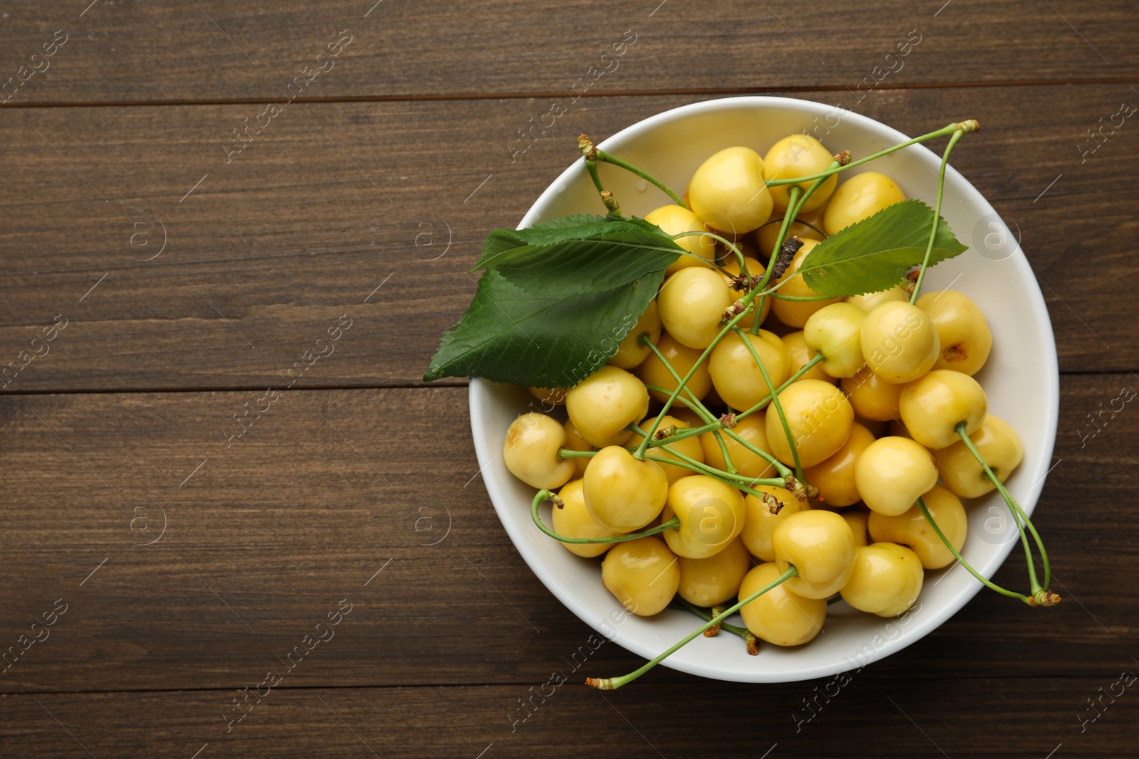 Photo of Bowl with ripe yellow cherries and green leaves on wooden table, top view. Space for text