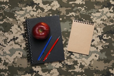 Photo of Notebooks, apple and pens on camouflage fabric, flat lay. Military education