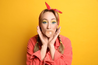 Photo of Fashionable young woman with braids and bright makeup blowing bubblegum on yellow background