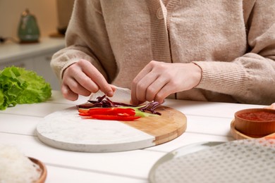 Photo of Making delicious spring rolls. Woman wrapping fresh vegetables into rice paper at white wooden table, closeup