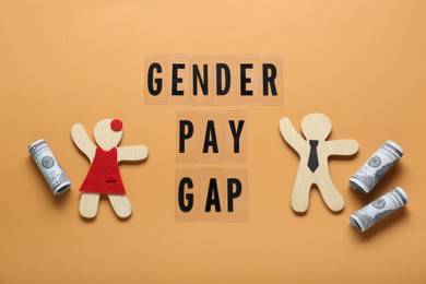 Gender pay gap. Wooden figures of man and woman with dollar banknotes on pale orange background, flat lay