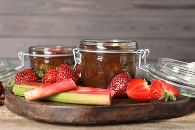 Jars of tasty rhubarb jam, fresh stems and strawberries on wooden table