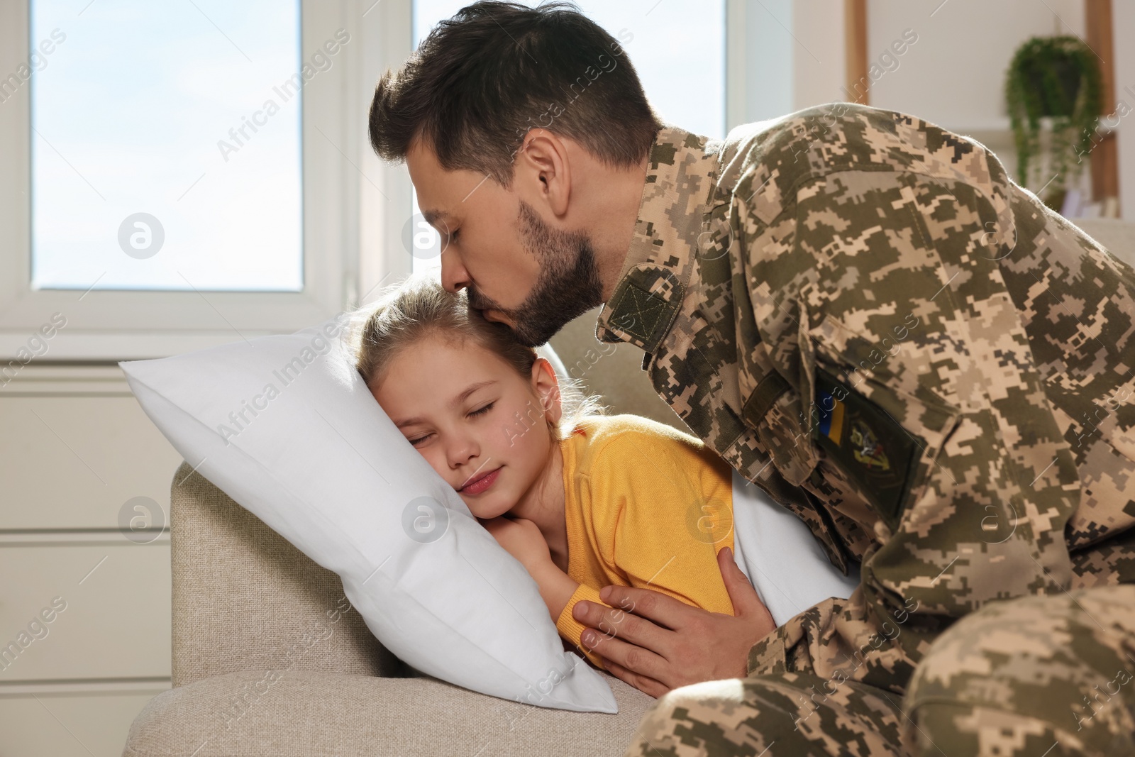 Photo of Soldier in Ukrainian military uniform kissing his daughter while she sleeping on sofa at home. Family reunion