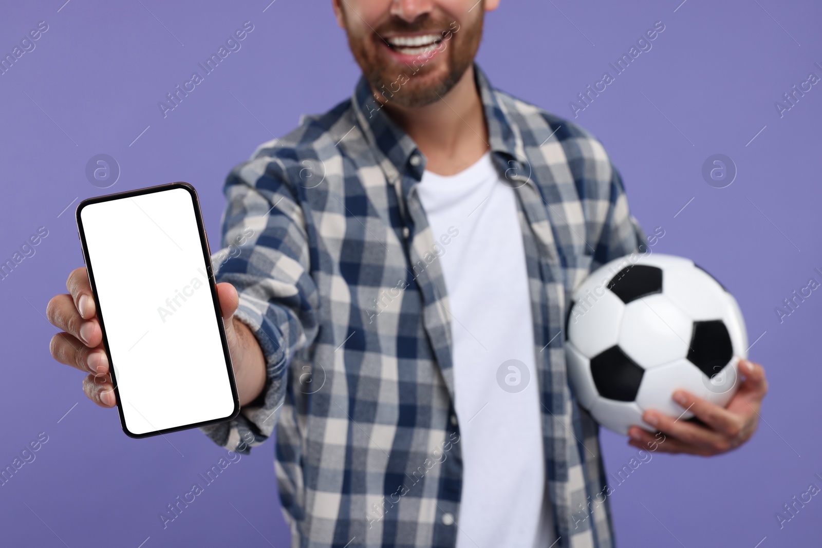 Photo of Happy sports fan with soccer ball and smartphone on purple background, closeup