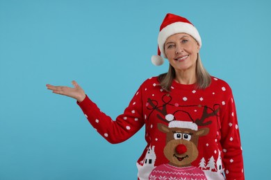 Happy senior woman in Christmas sweater and Santa hat showing something on light blue background. Space for text