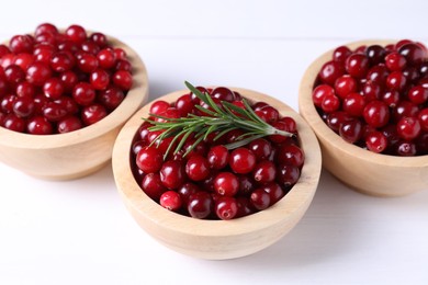 Photo of Fresh ripe cranberries in bowls and rosemary on white wooden table, closeup