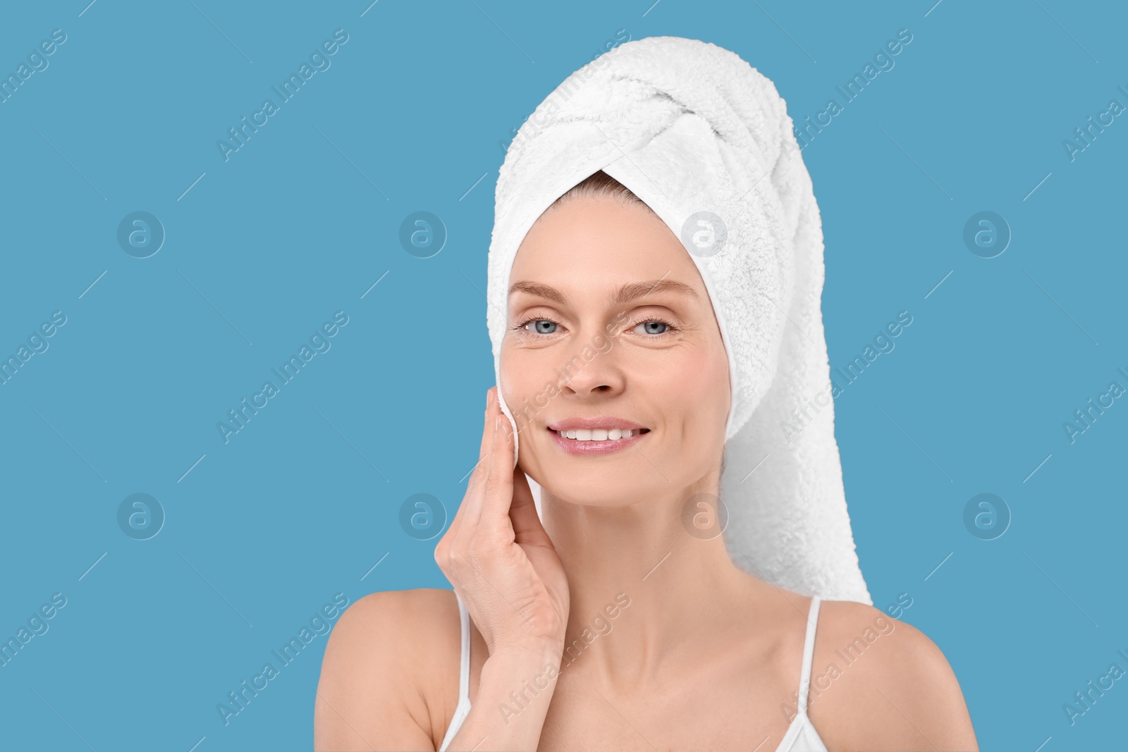 Photo of Beautiful woman in terry towel removing makeup with cotton pad on light blue background, space for text