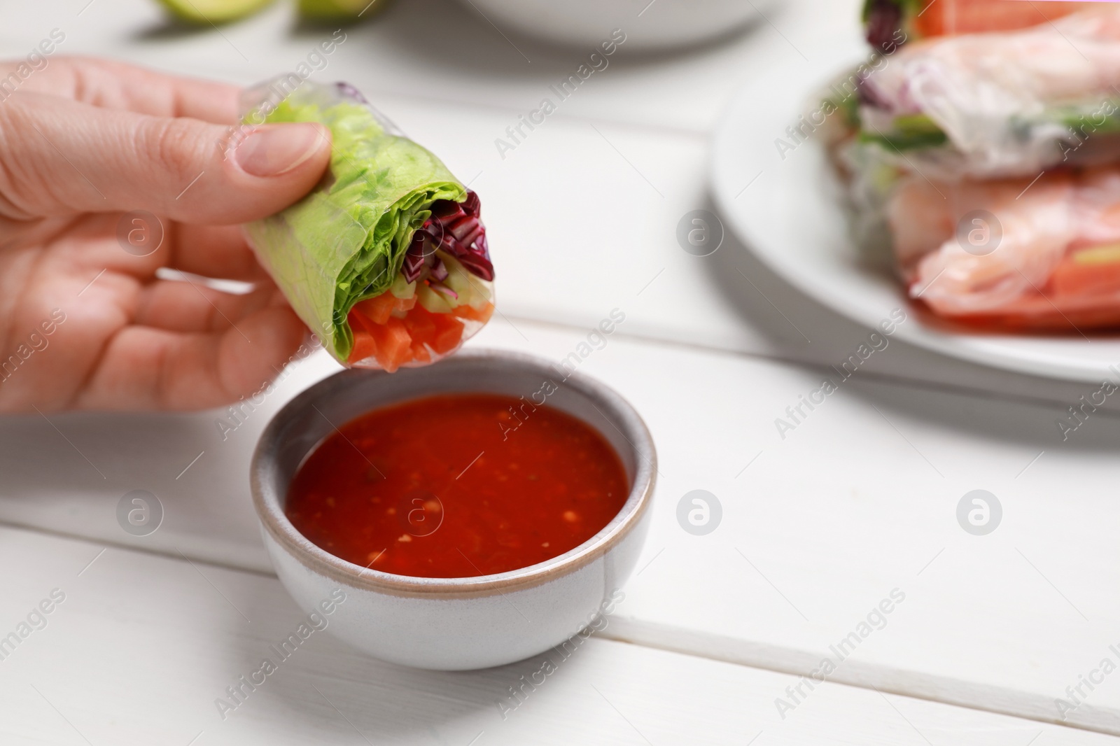 Photo of Woman eating delicious spring roll wrapped in rice paper with sauce at white wooden table, closeup