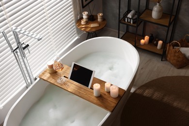 Wooden tray with tablet, wine and candles on bathtub in bathroom