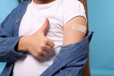 Photo of Girl with sticking plaster on arm after vaccination showing thumbs up against light blue background, closeup