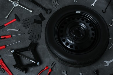 Car wheel, scissor jack and different tools on dark grey surface, flat lay