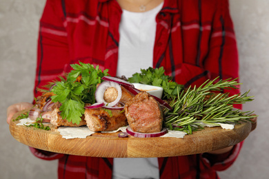 Photo of Woman holding wooden board with delicious roasted meat and herbal on grey background, closeup