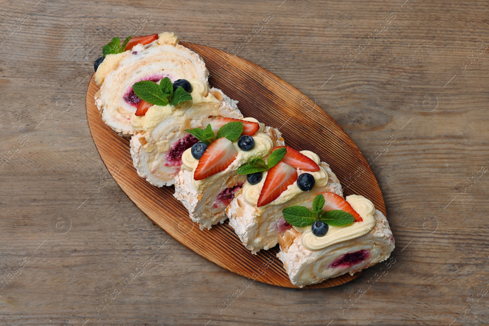 Photo of Pieces of tasty meringue roll with jam, cream, strawberry, blueberry and mint on wooden table, top view