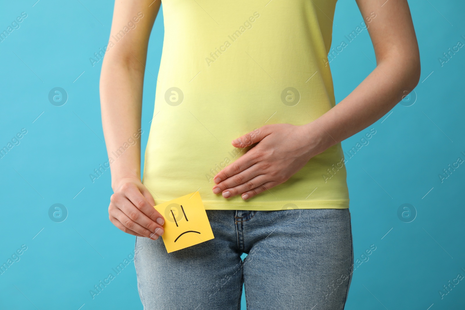 Photo of Woman holding sticky note with drawn sad face and suffering from cystitis on light blue background, closeup