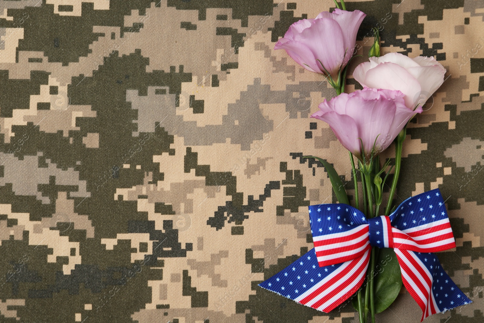 Photo of Top view of flowers and ribbon bow with American flag pattern on camouflage background, space for text. Armed Forces Day