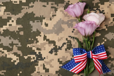 Photo of Top view of flowers and ribbon bow with American flag pattern on camouflage background, space for text. Armed Forces Day