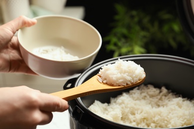 Woman putting tasty rice into bowl from cooker in kitchen, closeup