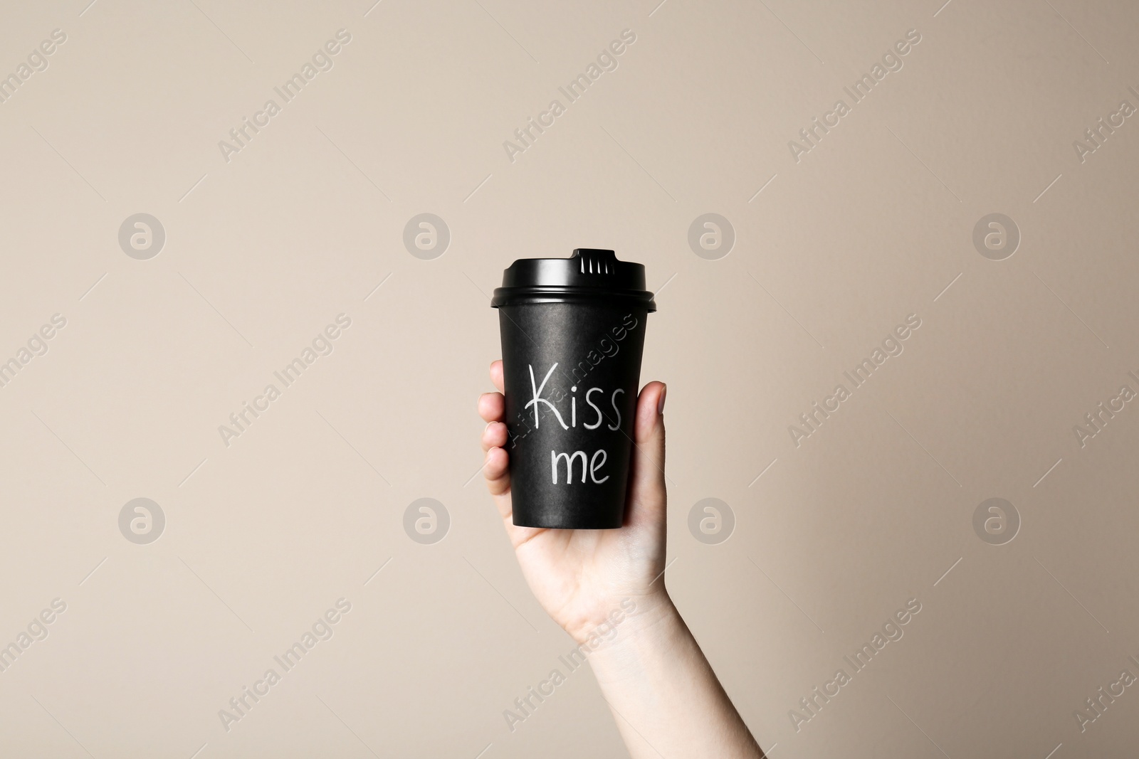 Photo of Woman holding paper cup with phrase Kiss Me on beige background, closeup