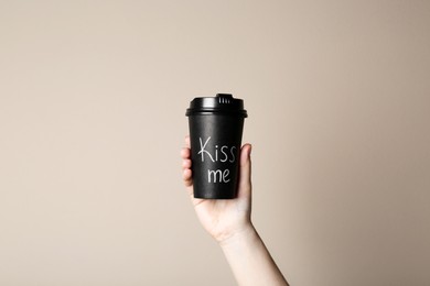Woman holding paper cup with phrase Kiss Me on beige background, closeup
