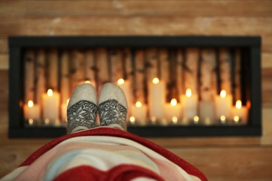 Photo of Woman wearing warm knitted socks near decorative fireplace indoors, closeup. Winter atmosphere