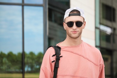 Photo of Handsome young man with stylish sunglasses and backpack near building outdoors, space for text