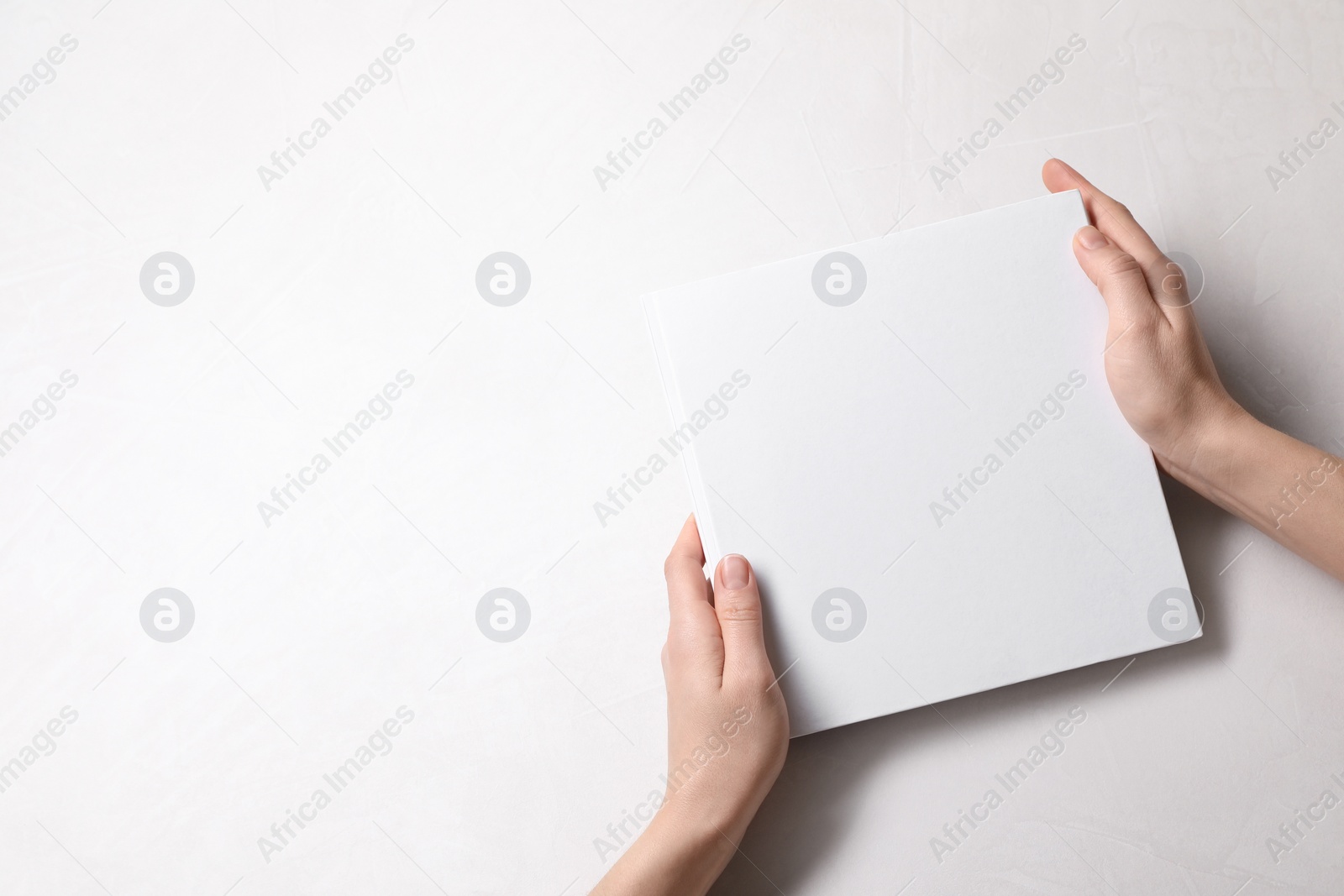 Photo of Woman holding book with blank cover at white table, top view. Mockup for design